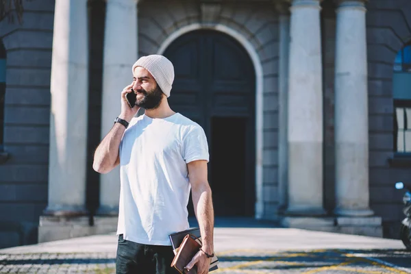 Handsome smiling creative male blogger having funny mobile conversation with friends via application on modern telephone.Happy bearded hipster guy talking on smartphone while walking at street