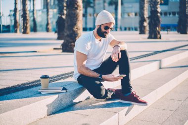 Hipster adam sakallı ve güneş gözlüğü güncelleştirme profili kullanarak 4 g Smartphone cep telefonu ile sosyal ağlar ile Internet. Arkadaşına kahve ile sokakta oturan cep telefonuna mesaj gönderme şapkalı şık öğrenci