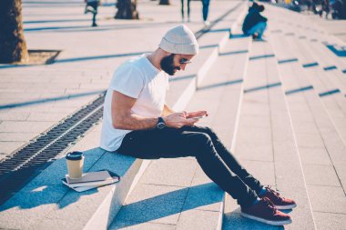 Şık genç adam sakallı ve merdiven outdooors kahve ile oturan Smartphone uygulama yüklenirken sosyal ağlarda takipçileri ile sohbet online Güneş gözlüğü ile gitmek için yan görünüm