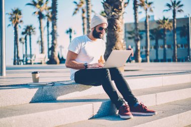 Neşeli sakallı blogger takipçileri ile dizüstü bilgisayar açık havada merdivenlerde oturan kendi blogunuzda yorum okuma. E-posta yolu ile 4 g netbook üzerinde kontrol güneş gözlüklü erkek grafik tasarımcı internet