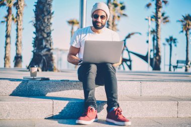 Dalgın sakallı serbest meslek sahibi netbook çalışma bağlı 4 g internet kentsel ortamda oturuyor. Gitmek için grafik tasarımcı kahve ile merdivenlerde dinlenme dizüstü bilgisayar video izleme güneş gözlüğü