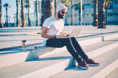 Sakallı öğrenci ders modern netbook üzerinde güneş gözlüğü bağlı kablosuz 4 g için açık havada gitmek için Internet kahve ile oturuyor. Şık erkek Tasarımcısı laptop yeni grafiği oluşturma