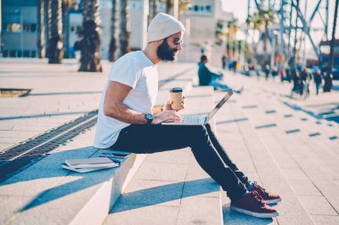 Sakallı hippi adam kahve elinde tutan ve kablosuz internet açık havada dinlenmek için bağlı dizüstü bilgisayarda internet sitelerinde son haberler okuma şık güneş gözlüğü yan görünüm