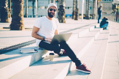 Neşeli portre yeni proje üzerinde dizüstü bilgisayar çalışma serbest geliştirirken kameraya gülümseyen güneş gözlüğü grafik tasarımcı sakallı. Pozitif hipster adam profil netbook üzerinde güncelleştirme