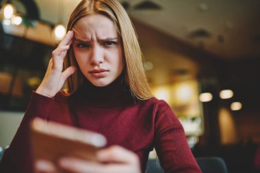 Half length portrait of charming sad blonde female holding hand next to head while working in coffee shop using gadget.Cropped image of tired hipster girl with painful headache looking at camera clipart