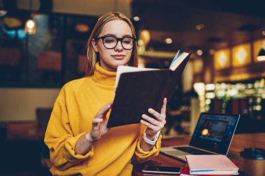 Genç çekici pozitif kadın öğrenci sınava hazırlık sırasında ilginç edebiyat okuma göz düzeltme için gözlük. Not defteri bilgileri analiz şirin akıllı hippi kız