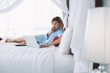 Pozitif genç kadın cep telefonu üzerinde Otel Oda Servisi rezervasyon yatakta, rahatlatıcı yapma ile kadın konuşurken telefon konuşmayı dizüstü bilgisayarda tatil sırasında göz haber okuma