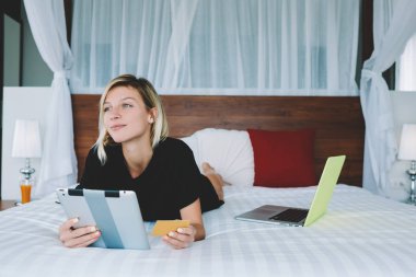 Çekici hippi kız taşınabilir pc satın alma kredi kartı ödeme ve e-ticaret için tutarak web mağaza satın alma hakkında düşünme genç kadın evde, rahat yatak üzerinde rahatlatıcı kullanarak çevrimiçi alışveriş yapma