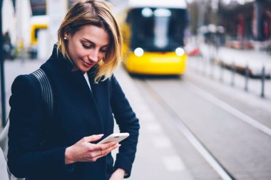 Gülümseyen kadın turist üzerinde durmak tramvaylar City, pozitif kadın ulaşım bilgileri web sitesi hücresel zamanında alma konusunda göz tarih memnun olarak trafik yolları kontrol smartphone ile ayakta