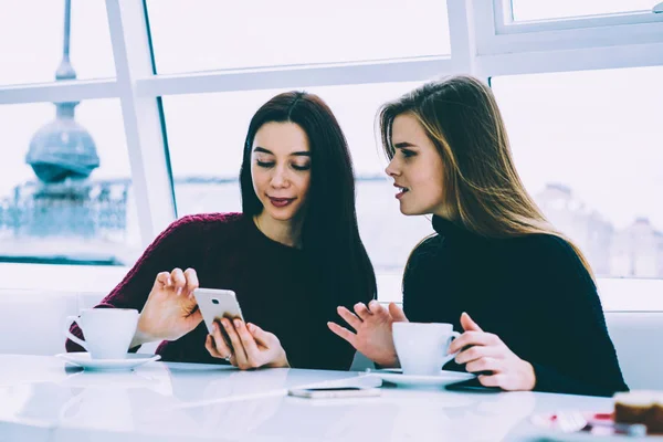 Charming experienced blonde entrepreneur explaining to brunette colleague general ideas of business project while searching useful information in web pages on cellphone connected to 4G indoors