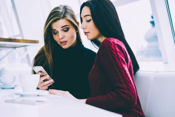 Pondering attractive female friends carefully watching interesting video on cellular connected to 4G internet.Gorgeous young students reading news in websites while recreating in loft cafeteria