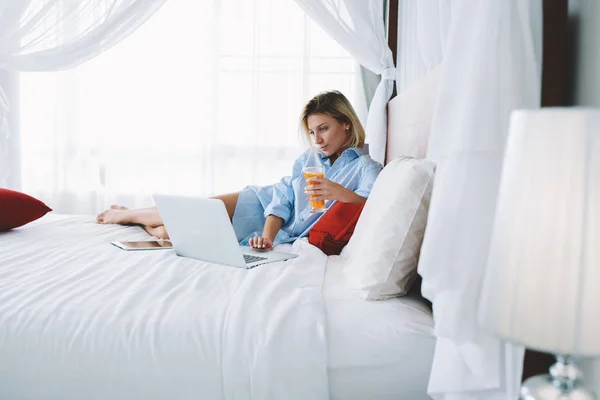 在笔记本电脑上看电影的年轻女子上午在床上放松与新鲜的果汁 女性自由职业者享受远程工作 使用上网本连接到 Wifi 为阅读新闻在酒店房间重新创建 — 图库照片