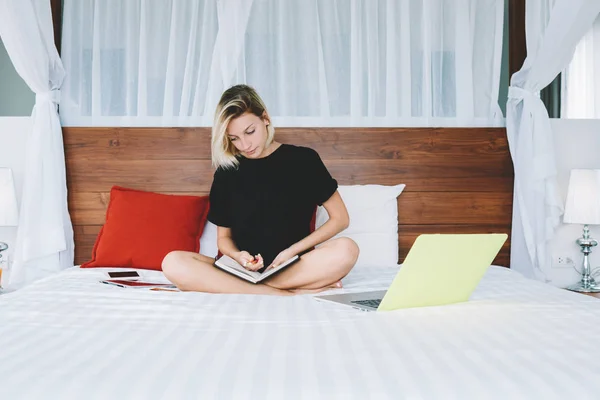 Pensive female student sitting at bed with laptop computer and textbook doing homework task, smart young woman spending time on autodidact at home interior  learning information prepare for exams
