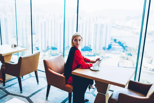 Serious businesswoman looking away holding smartphone for checking notification sitting at table near window in cafe,female owner waiting for meeting on coffee break holding cellular for contact
