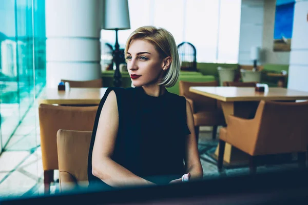 Gorgeous businesswoman with makeup and red lipstick dressed in elegant outfit looking away spending time in cafe interior, prosperous blonde female owner in formal wear thinking about future plans