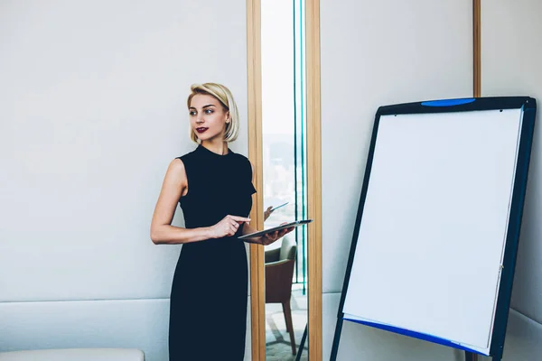 Gorgeous female leader of company standing near flip chart with touchpad making presentation, prosperous business woman in elegant formal outfit making speech on economic conference using tablet