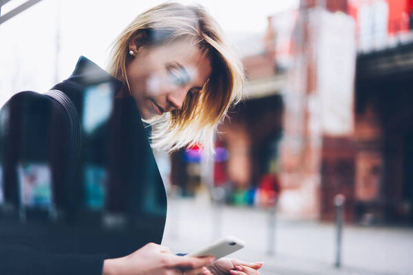 Blonde female tourist using smartphone traffic online app for monitoring news about bus departure, thoughtful hipster girl read information via cellular waiting for transport on stop in downtown