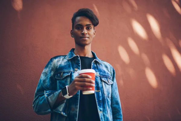 Half length portrait of hindu young man with coffee to go looking at camera.Serious international teenager in denim jacket standing against promotional background for your advertising text message
