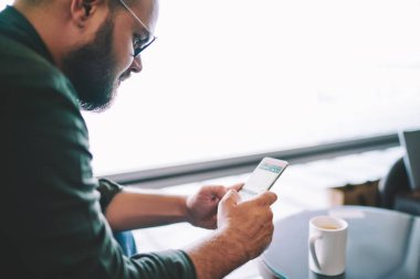 Kırpılmış görüntü bir kafede çay bir fincan ile oturan smartphone ücretsiz yüksek hızlı internet bağlantısı ile çevrimiçi peyment yapma Eyewear dalgın sakallı adam. Blogger paylaşım medya hücresel ağları