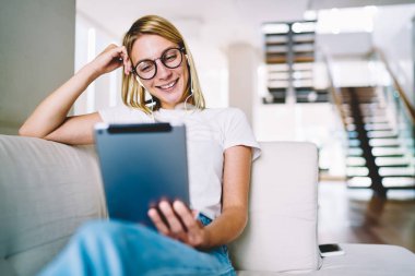 Optik gözlükler komik eğitsel bir dokunmatik yüzey evde kanepede oturan izlerken pozitif sarışın genç kadın. Kulaklık takipçileri Tablet yorum okuma sırasında gülüyor içinde neşeli blogger