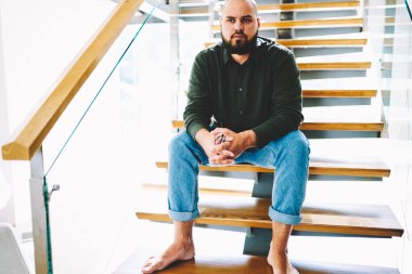 Düşünceli sakallı genç adam elinde gözlük ile ahşap merdivenlerde oturan ve hafta sonu eğlence süre boyunca kendi daire iç üzerinde düşünme şık casual kıyafet giymiş