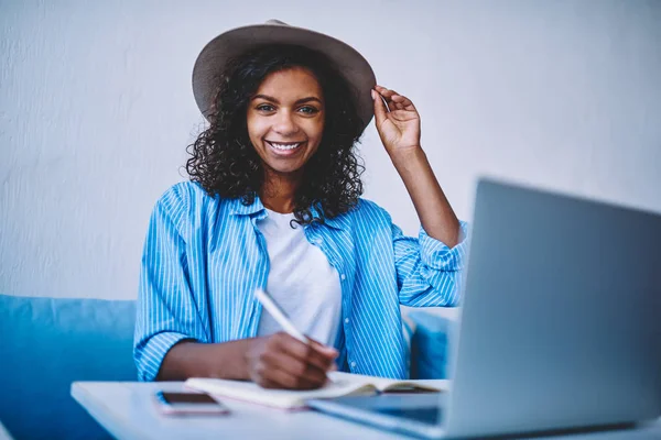 Portrait of afro american female freelancer with curly hair and hat making plan of project earning money online,smiling hipster girl looking at camera writing in notepad learning via online courses