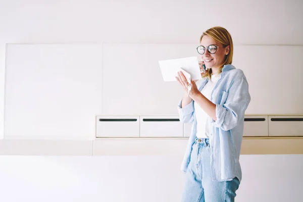 Happy blonde young woman in eyeglasses received letter with good news about winning discounts in webstore and calling to internet consultant to check information on smartphone standing near mailbox