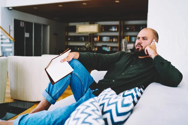 Bearded young man having mobile conversation on smartphone device while spending leisure time on reading book at home in weekend.Male communicating on cellular resting with textbook on cozy couch