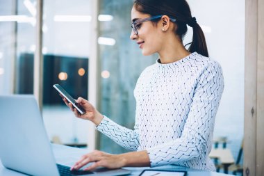 Bilgi e-posta gönderme telefonla sohbet kadın idari işler müdürü gülümseyen, işgal okuma mesaj smartphone ayakta ofiste masa üzerinde pozitif resepsiyonist memnun
