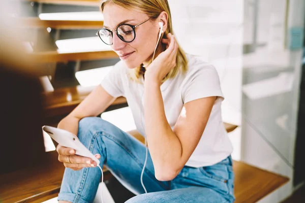 Cropped image of young attractive happy blonde woman in trendy eyeglasses spending leisure time at home with music. Charming positive hipster girl enjoying resting while listening to cool playlist