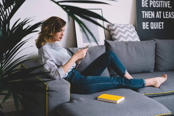 若い女性ブロガーは スタイリッシュなアパートで快適なソファに横たわってスマートフォン上の無線ホームインターネットを介してウェブサイト上で新しい投稿を公開します — ストック写真