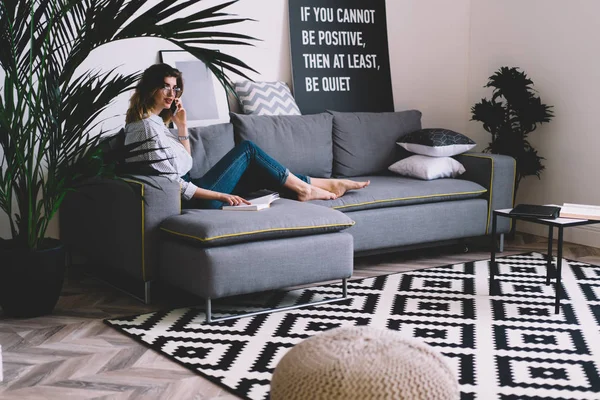 Portrait of attractive hipster girl communicating with friend on smartphone while read book and resting on comfortable sofa in fashionable home interior.Stylish blogger talking on mobile phone