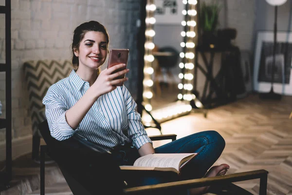 Positive young woman resting with book on chair at home stylish interior while reading notification with good news on smartphone.Cheerful female blogger watching funny video in blog enjoying free time