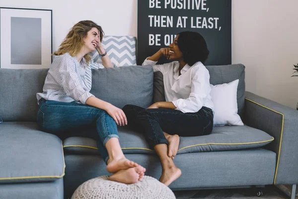Cheerful african american young woman laughing together with best caucasian friend during funny conversation.Two positive women talking about funny story and relaxing on couch in stylish home interior