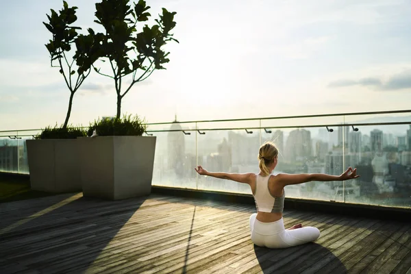 Sports woman in activewear sitting in pose during spiritual practice on rooftop restore vitality and get inspire, back view slim girl with perfect body shape having yoga workout on building terrace