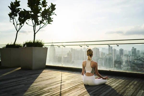 Caucasian woman in active wear meditating in lotus pose feeling zen on top of building in city, back view of sportive girl having yoga training for keeping perfect body and vitality in megalopolis