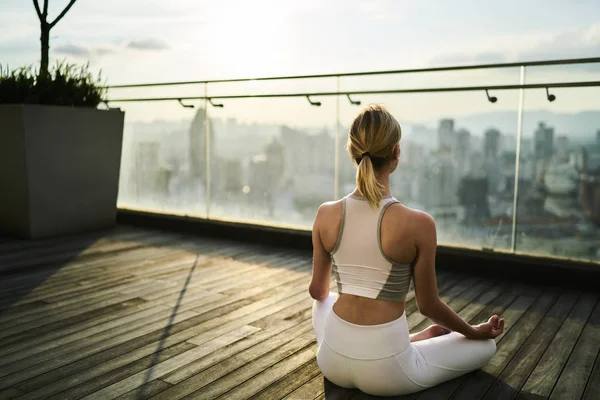 Sports woman in white track suit sitting in lotus pose meditating and feeling calm in big city, back view of slim woman with perfect figure having yoga training on building rooftop relaxing in morning