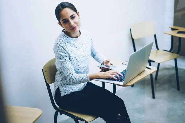 Young woman spending time in college classroom using wireless connection for browsing education web page on laptop computer, female student waiting for lesson begins sitting at desk updating software