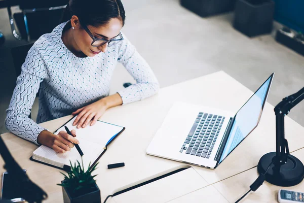 Concentrated female manager pondering on idea for publication writing in notepad before upload on laptop computer, pensive business woman planning statement of startup spending time at desktop