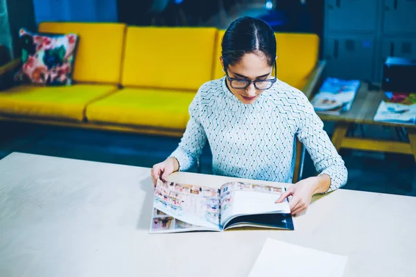 Pensive female editor of magazine reading issue for checking articles and publication while sitting at table ,concentrated woman choosing items in paper catalogue for buying furniture in office