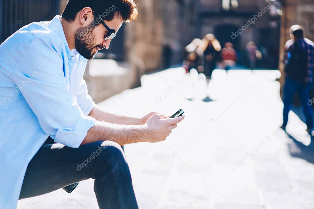 Side view of casual dressed male blogger in sunglasses typing text on smartphone fir publication on website sitting in downtown.Young man chatting with friends online via 4 internet connection
