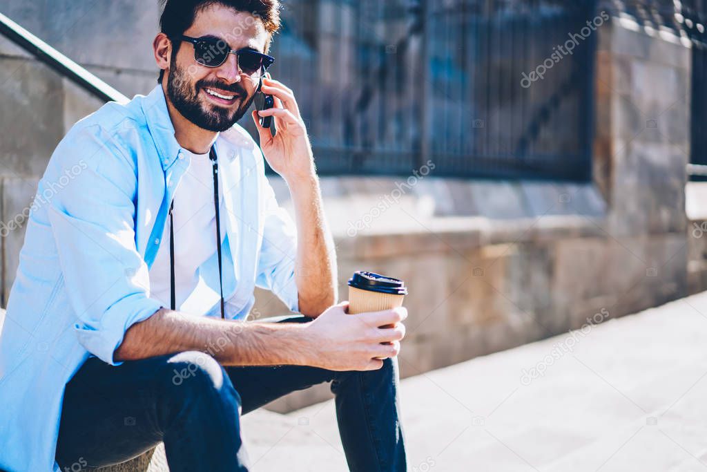 Portrait of happy bearded hipster guy in stylish sunglasses smiling at camera while having funny mobile conversation with friend in roaming on smartphone sittting on street with coffee to go in hand