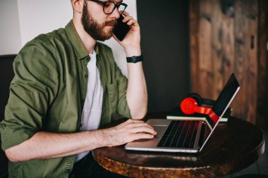 Şık sakallı hippi erkek tarafından smartphone kız arkadaşıyla konuşuyor ve uzay, genç profesyonel programcı adam arayan müşteri site oluşturma hakkında ayrıntılı bilgi tartışmak için coworking içinde dizüstü bilgisayar kullanarak