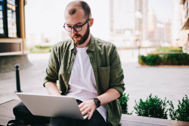 Dizüstü bilgisayar ile bankta oturmuş ve yeni modern elektronik aygıt için Gözat Web sitesi kullanarak proje hazırlama webinar online, profesyonel programcı izlerken kendinden emin ve düşünceli genç adam
