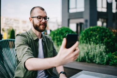 Trendy erkek holding cep telefonu cep telefonu kamera ve kendini açık havada kafede oturan ve selfie fotoğrafı tıklatın smartphone kullanarak beyaz hipster adam alarak fotoğraf gülümseyen elinde sakallı