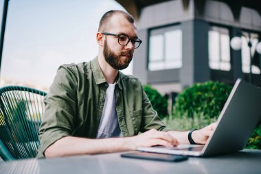 Sakallı yakışıklı modern elektronik cihaz, film izlerken konsantre açık havada oturma ve çevrimiçi eğitim için online webinar izlerken yeni dizüstü bilgisayar kullanan genç beyaz hipster adam