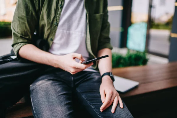 Cropped photo of hipster guy sitting outside and trying check mail by smartphone, male chatting with friends using new modern mobile phone while watching video online and reading notification