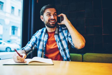 Pozitif sakallı hippi adam smartphone aygıt üzerindeki telefon görüşmesi sırasında gülüyor. Uzay coworking oturan telefonda konuşurken Not Defteri'nde bilgi yazma rahat kıyafet içinde neşeli erkek blogger