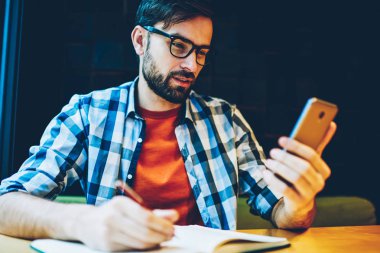 Dalgın sakallı öğrenci Not defteri smartphone aygıtından metin bilgileri yeniden yazma. Web sitesinde telefon haber okuma ve kafede oturan Not defterinizdeki notlar yapma gözlük hipster adam