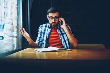 Ders kitabı tahta masada oturan dijital smartphone aygıt arama gözlük şok sakallı genç adam. Şaşırmış gibi yenilikçi öğrenci modern telefonla konuşurken rahat kıyafet giymiş